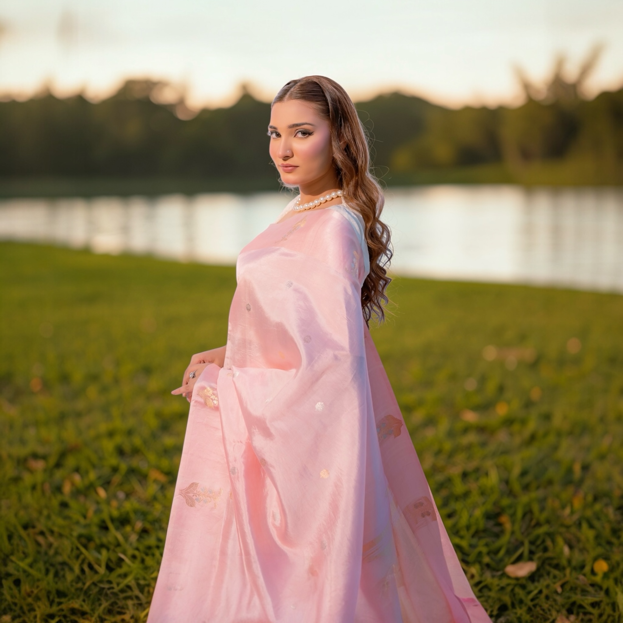 Woman in a Baby pink Puna silk Banarasi saree with sona rupa buti motifs standing in a grassy field with a lake in the background