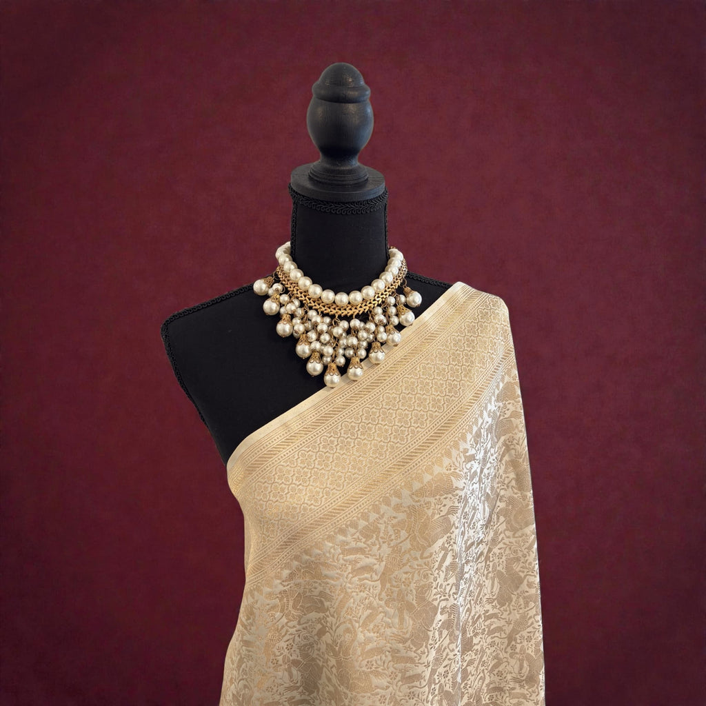Mannequin wearing a off-white handwoven banarasi saree with a gold necklace against a maroon background