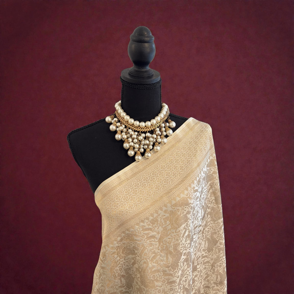 Mannequin wearing a off-white handwoven banarasi saree with a gold necklace against a maroon background