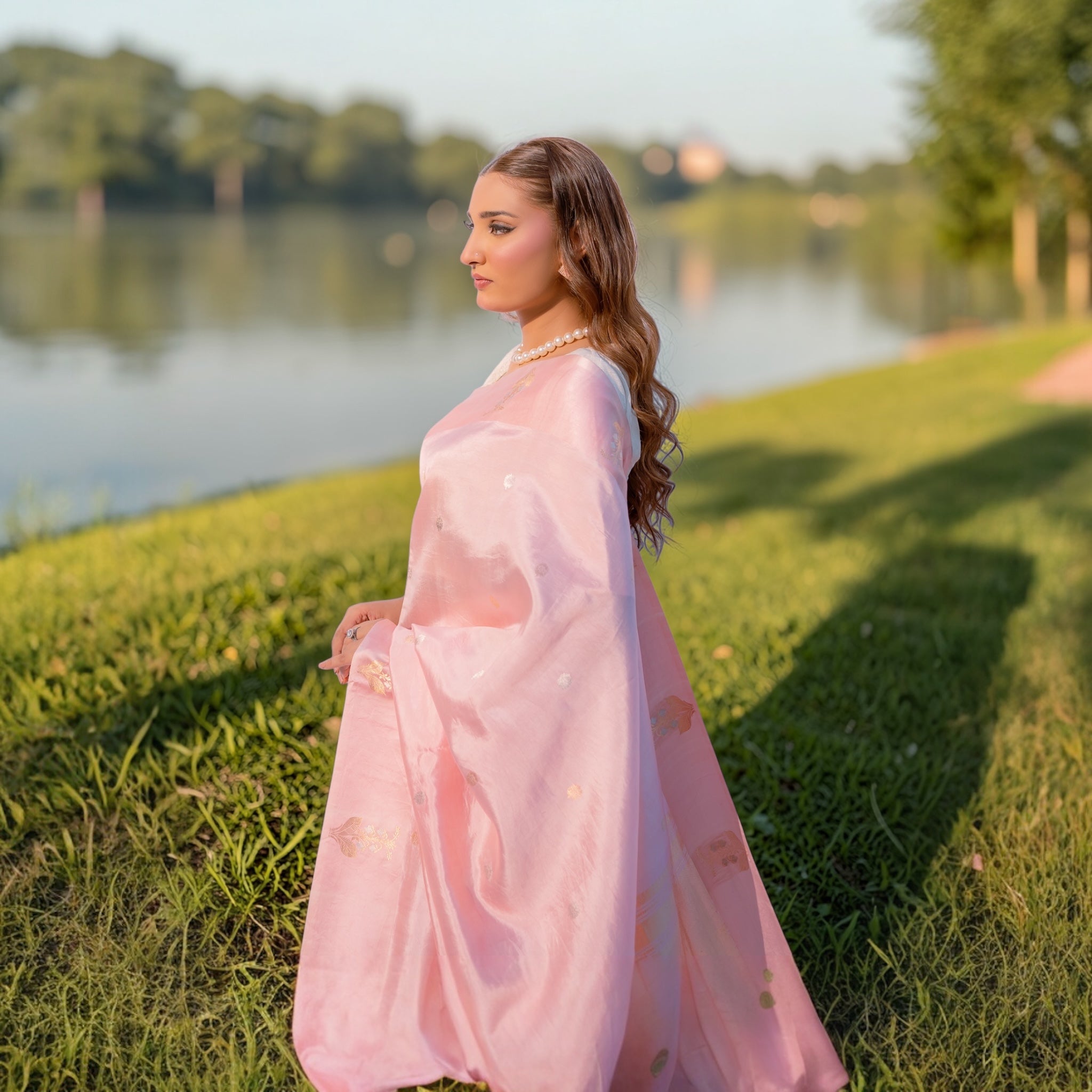 A woman wearing the Sacred Hues 1.2 Baby Pink Banarasi Saree in Puna silk with gold and silver Sona Rupa motifs, standing in a green field by a lake at sunset.