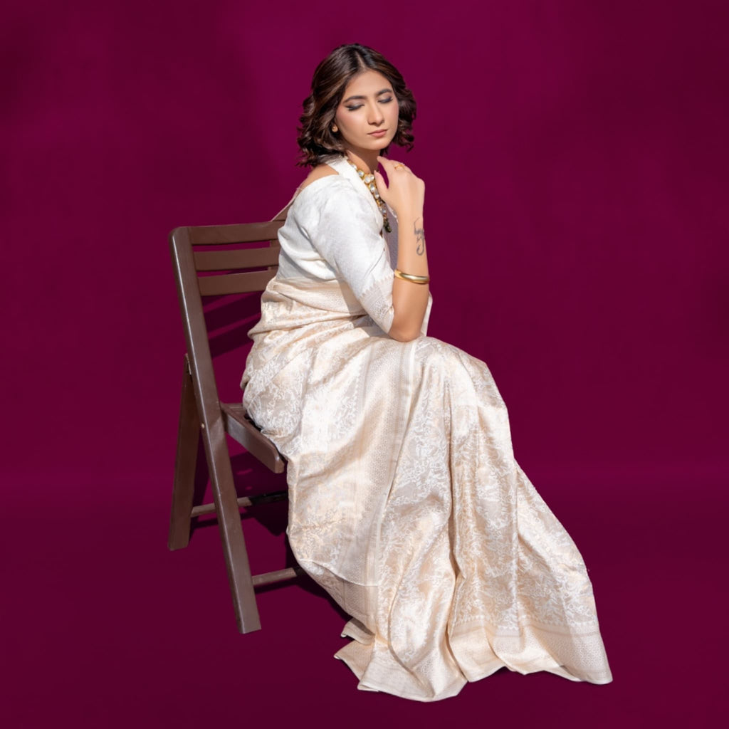 Woman in a handwoven off-white banarasi saree sitting on a wooden chair against a purple background