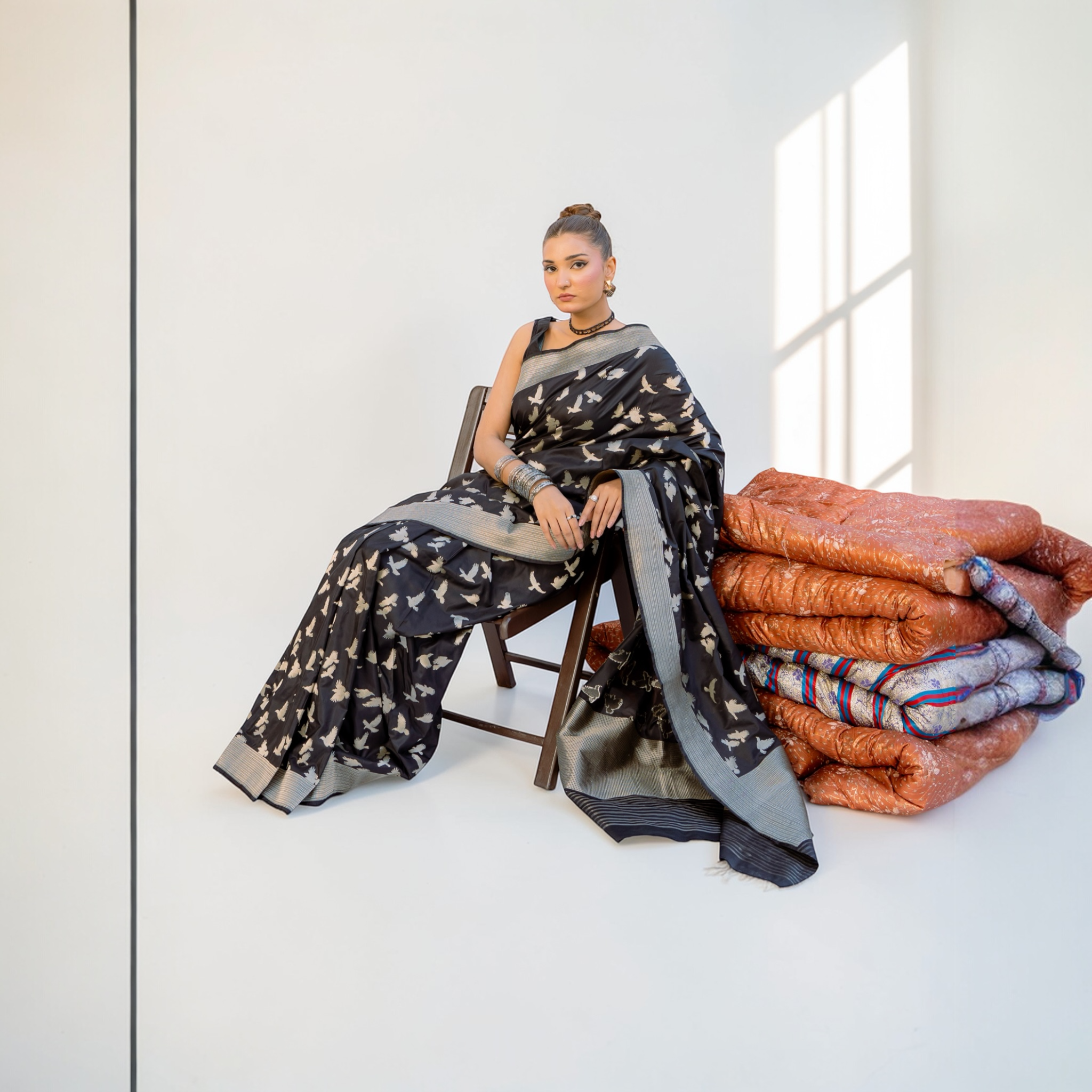 Woman sitting on a chair wearing a black banarasi saree with a stack of folded clothes next to her - Lunarae London