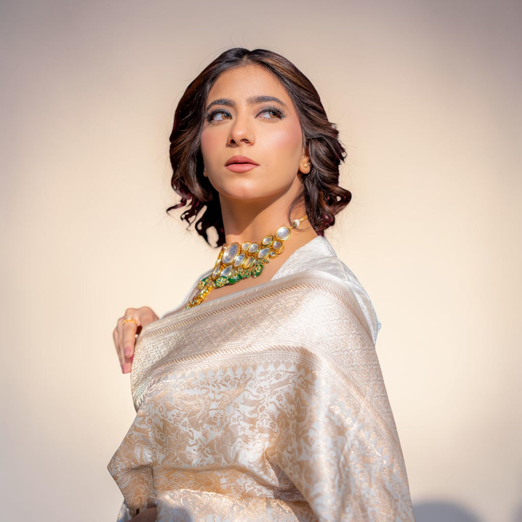 Woman wearing a white saree with gold jewelry against a beige background