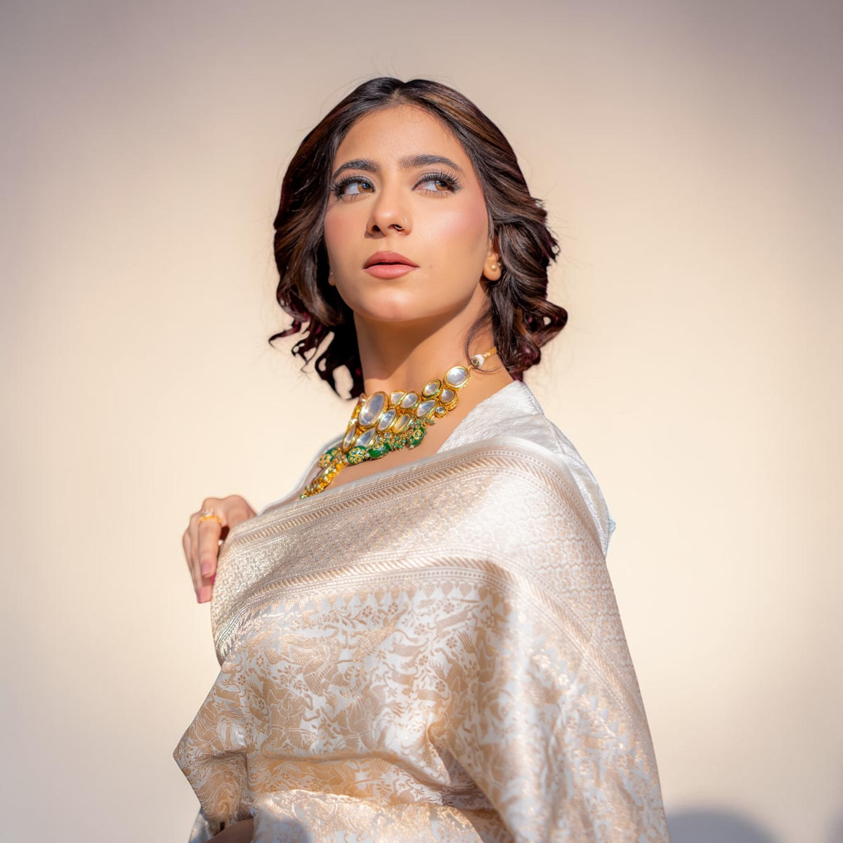 Woman wearing a white saree with gold jewelry against a beige background