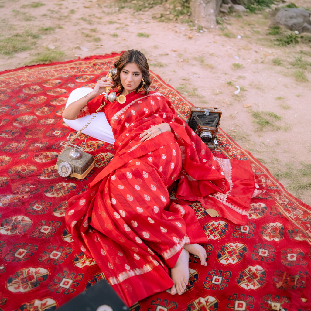 Woman reclining on a red rug wearing a Sacred Hues 1.1 Red Banarasi Silk Saree with vintage telephone and camera props.