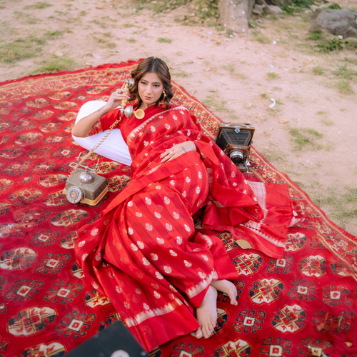 Woman reclining on a red rug wearing a Sacred Hues 1.1 Red Banarasi Silk Saree with vintage telephone and camera props.