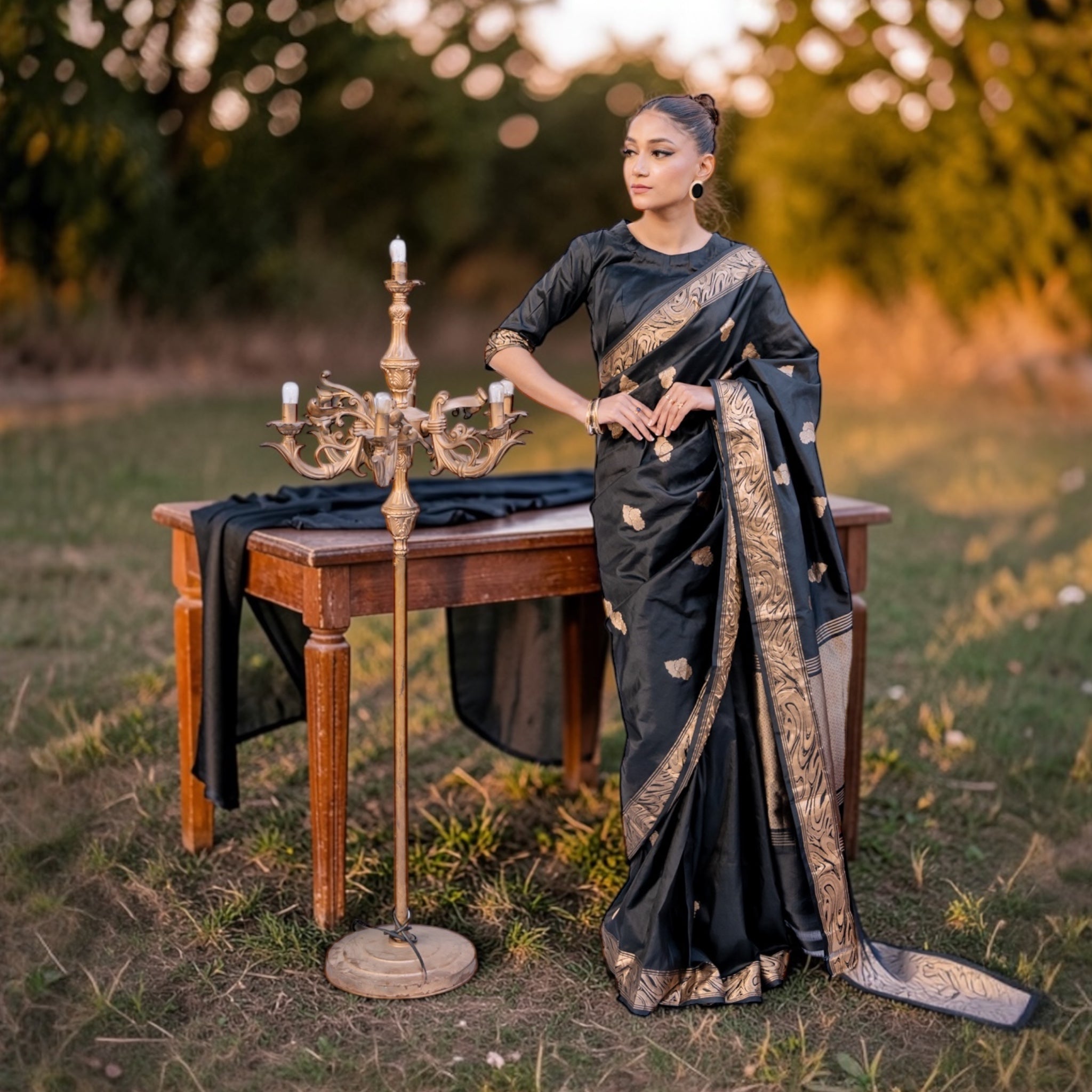 Women in a Black Pure Katan silk Banarasi saree with golden floral motifs from Sacred Ties collection standing next to a wooden table outdoors.